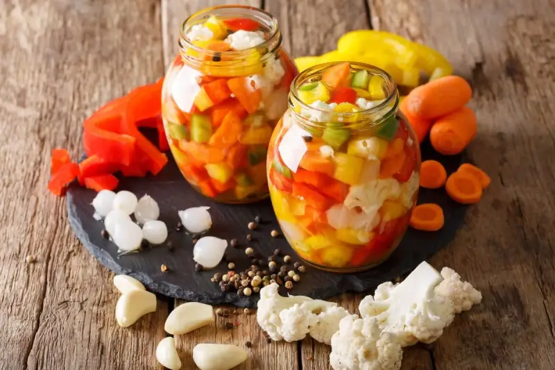 Due barattoli di giardiniera fatta in casa, pieni di verdure colorate come peperoni, carote e cavolfiore, su un tagliere di ardesia con ingredienti freschi intorno.